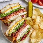 Chicken Club Sandwich cut into halves on a white plate, layered with lettuce, tomato, bacon, cheddar cheese, and sliced chicken breast, served with potato chips and a dill pickle, close-up, top-down view, vibrant and fresh lunch presentation.