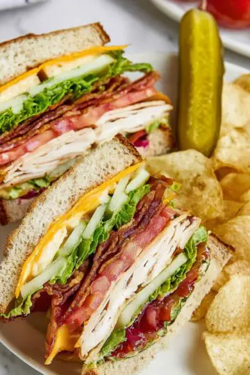 Easy Chicken Club Sandwich Recipe 1 Chicken Club Sandwich cut into halves on a white plate, layered with lettuce, tomato, bacon, cheddar cheese, and sliced chicken breast, served with potato chips and a dill pickle, close-up, top-down view, vibrant and fresh lunch presentation.