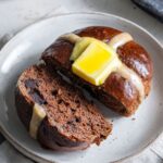 Chocolate Hot Cross Buns A chocolate hot cross bun sliced open on a white plate with a slab of butter melting inside, next to a cup of tea and a butter knife.