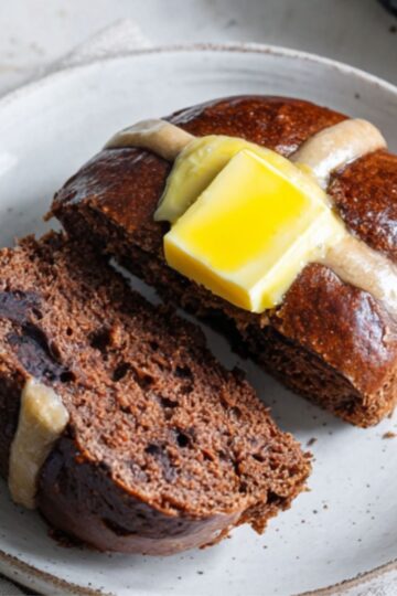 Healthy Chocolate Hot Cross Buns Recipe 1 Chocolate Hot Cross Buns A chocolate hot cross bun sliced open on a white plate with a slab of butter melting inside, next to a cup of tea and a butter knife.