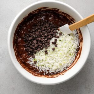 Best Chocolate Zucchini Bread Recipe 15 A white mixing bowl with chocolate zucchini bread batter topped with shredded coconut and chocolate chips, with a spatula resting inside.