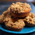 Coffee Cake Donuts Close-up of freshly baked coffee cake donuts stacked on a vibrant blue plate, featuring golden-brown crumbly streusel topping and soft, moist interior. Warm natural kitchen lighting highlights the texture, creating a cozy, homemade bakery feel, ideal for food blogs, recipe tutorials, or Pinterest dessert posts