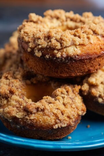 Healthy Coffee Cake Donuts Recipe 1 Coffee Cake Donuts Close-up of freshly baked coffee cake donuts stacked on a vibrant blue plate, featuring golden-brown crumbly streusel topping and soft, moist interior. Warm natural kitchen lighting highlights the texture, creating a cozy, homemade bakery feel, ideal for food blogs, recipe tutorials, or Pinterest dessert posts