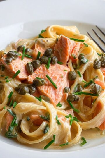 The Best Creamy Salmon Pasta Recipe 1 Creamy Salmon Pasta Creamy pasta with salmon pieces and capers, garnished with chopped chives, served on a white plate with the pasta pan in the background.