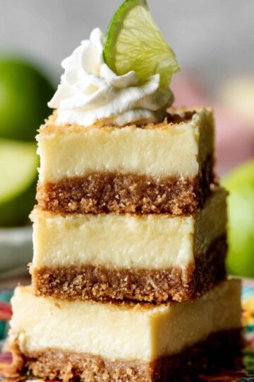 Healthy Key Lime Pie Bars Recipe 2 Key Lime Pie Bars Three square Key Lime Pie bars stacked on a colorful floral plate. Each bar has a thick, creamy pale-yellow filling over a crumbly brown crust. The top bar is garnished with a swirl of whipped cream and a small wedge of fresh lime. Whole limes are blurred in the background.