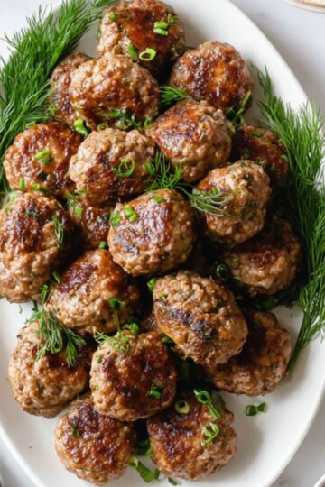 Best Homemade Kotleti Recipe 1 Kotleti Recipe A white oval platter filled with cooked meatballs garnished with fresh dill, surrounded by bowls of sauce, pickles, and a dish of grains, on a white table.
