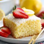 Lemon Buttermilk Cake A square slice of lemon cake on a white plate, garnished with fresh strawberries and lemon wedges, ready to serve.