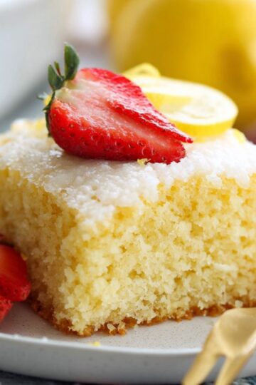 Healthy Lemon Buttermilk Cake Recipe 1 Lemon Buttermilk Cake A square slice of lemon cake on a white plate, garnished with fresh strawberries and lemon wedges, ready to serve.