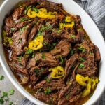 Mississippi Pot Roast Close-up of fully cooked Mississippi pot roast served in a white dish, garnished with parsley and pepperoncini peppers, with a gray-striped cloth nearby