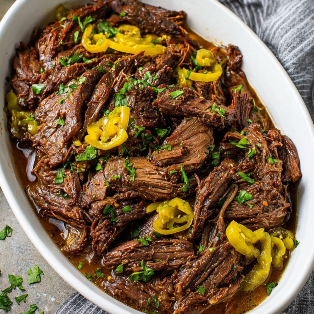 Mississippi Pot Roast Close-up of fully cooked Mississippi pot roast served in a white dish, garnished with parsley and pepperoncini peppers, with a gray-striped cloth nearby