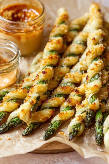 Easy Parmesan Asparagus Pastry Twists Recipe 2 Parmesan Asparagus Pastry Twists Golden-brown puff pastry twists wrapped around asparagus spears are arranged on parchment paper. Two small jars of dipping sauces, one reddish and one yellow-orange, sit next to the twists.