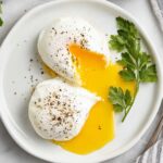 Poached Eggs Overhead view of two perfectly poached eggs on a white plate, one cut open with runny yolk, garnished with fresh parsley, with a striped kitchen towel, salt, and two whole eggs in the frame, bright natural lighting