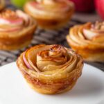 Puff Pastry Apple Roses A close-up of a single baked apple rose pastry on a white round stand, with several more apple roses cooling on a wire rack in the background and green and red apples nearby.