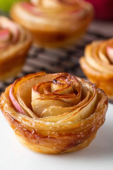 Delicious Puff Pastry Apple Roses Recipe 1 Puff Pastry Apple Roses A close-up of a single baked apple rose pastry on a white round stand, with several more apple roses cooling on a wire rack in the background and green and red apples nearby.