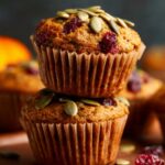 Pumpkin Cranberry Muffins A stack of pumpkin cranberry muffins topped with green pumpkin seeds, perfect for an autumn treat, with a background of seasonal pumpkins.