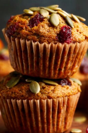 The Best Pumpkin Cranberry Muffins 1 Pumpkin Cranberry Muffins A stack of pumpkin cranberry muffins topped with green pumpkin seeds, perfect for an autumn treat, with a background of seasonal pumpkins.