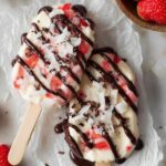 Strawberry Yogurt Clusters Four freshly poured strawberry yogurt popsicles on wooden sticks with diced strawberries visible, placed on white parchment paper before freezing
