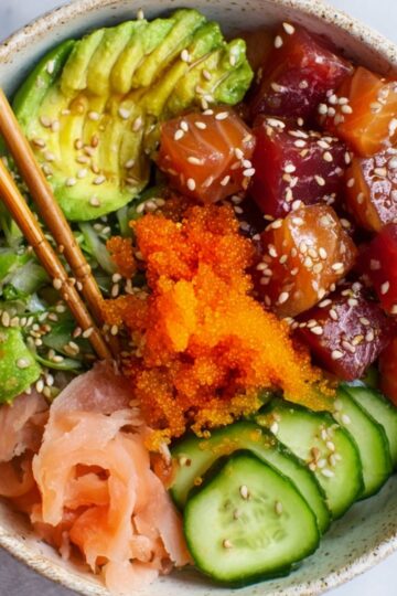 Delicious Sushi Poke Bowls Recipe 3 Sushi Poke Bowls A colorful poke bowl featuring cubed raw salmon and tuna, sliced avocado, cucumber, pickled ginger, and orange tobiko, garnished with sesame seeds, with a pair of chopsticks resting on the side of the bowl.