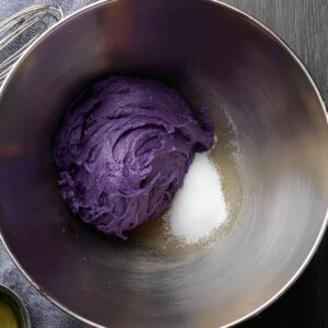The best Ube Crinkles Recipe 14 Metal mixing bowl with purple ube jam, oil, and sugar ready to be combined for cookie dough preparation, placed on a dark surface with a cloth napkin.