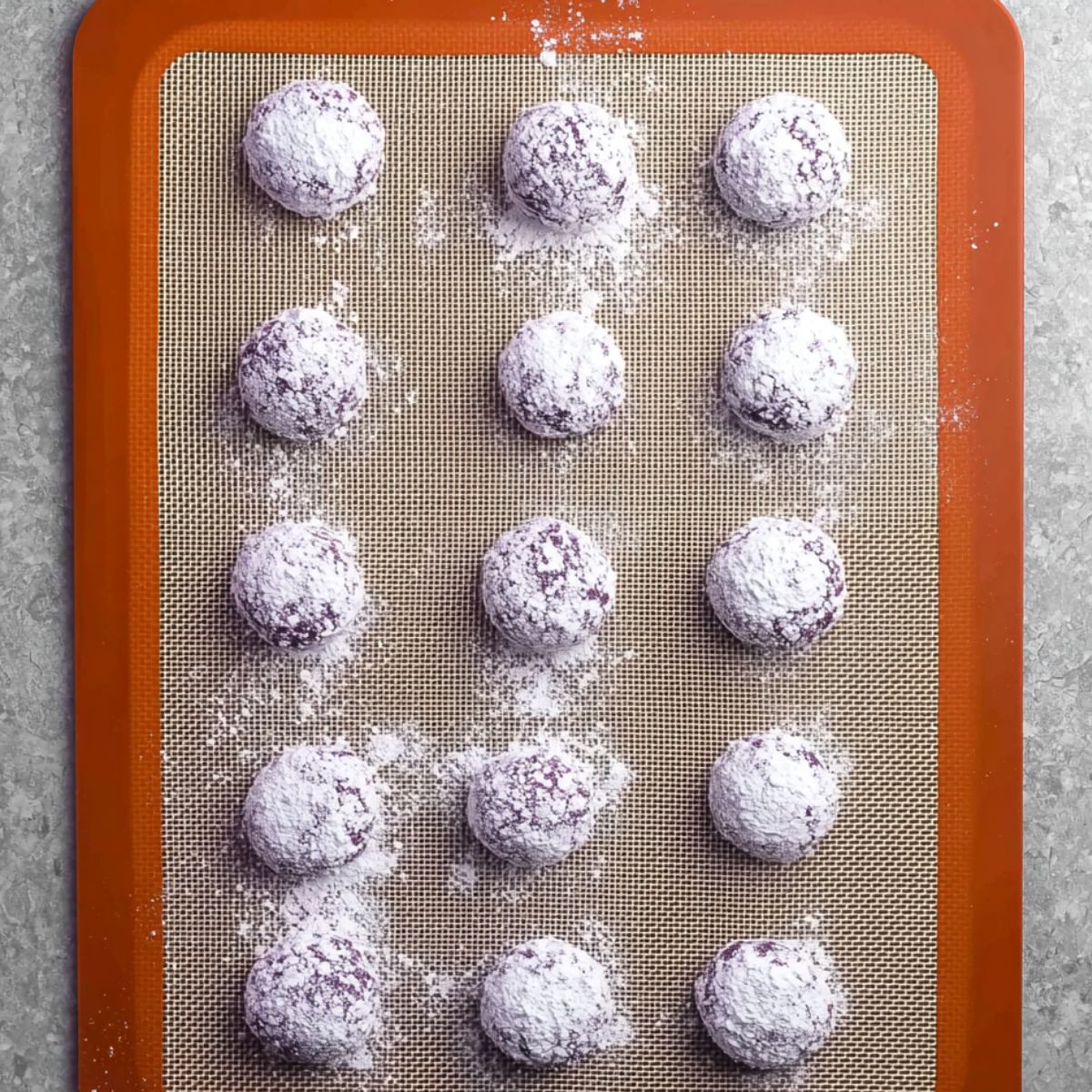 The best Ube Crinkles Recipe 12 Ube Crinkles Overhead view of a silicone baking mat with 12 round purple cookie dough balls coated in powdered sugar, evenly spaced and ready to bake.