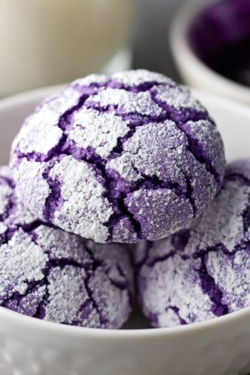 The best Ube Crinkles Recipe 3 Ube Crinkles Three baked purple ube crinkle cookies in a white bowl, coated with powdered sugar, with a glass of milk and a small bowl of ube jam blurred in the background.