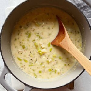 Close-up of creamy clam chowder in a gray pot with diced celery, clams, and potato pieces visible. A wooden spoon is dipped into the soup, highlighting the thick texture.