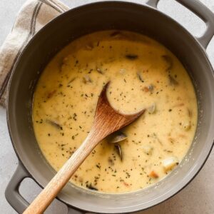A gray Dutch oven filled with creamy New England-style clam chowder. The soup has visible chunks of clams, potatoes, and herbs. A wooden spoon rests in the pot, partially submerged in the chowder.