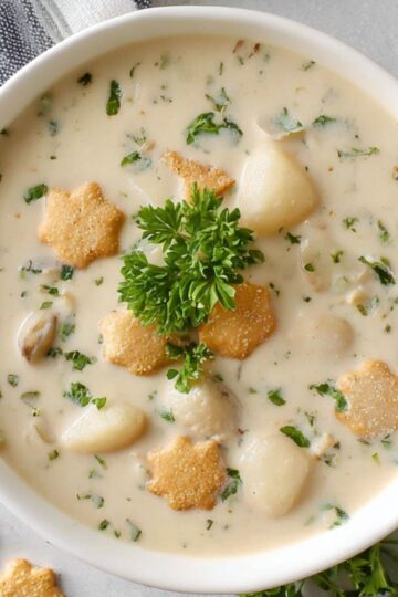 Easy Clam Chowder Recipe 1 clam chowder recipe A white bowl of creamy clam chowder garnished with parsley and oyster crackers in star and round shapes. The chowder is thick with chunks of potato, clams, and vegetables, served on a light gray surface.