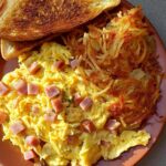 crispy hash browns A plate of scrambled eggs with diced ham, served with crispy hash browns and a slice of toasted bread.