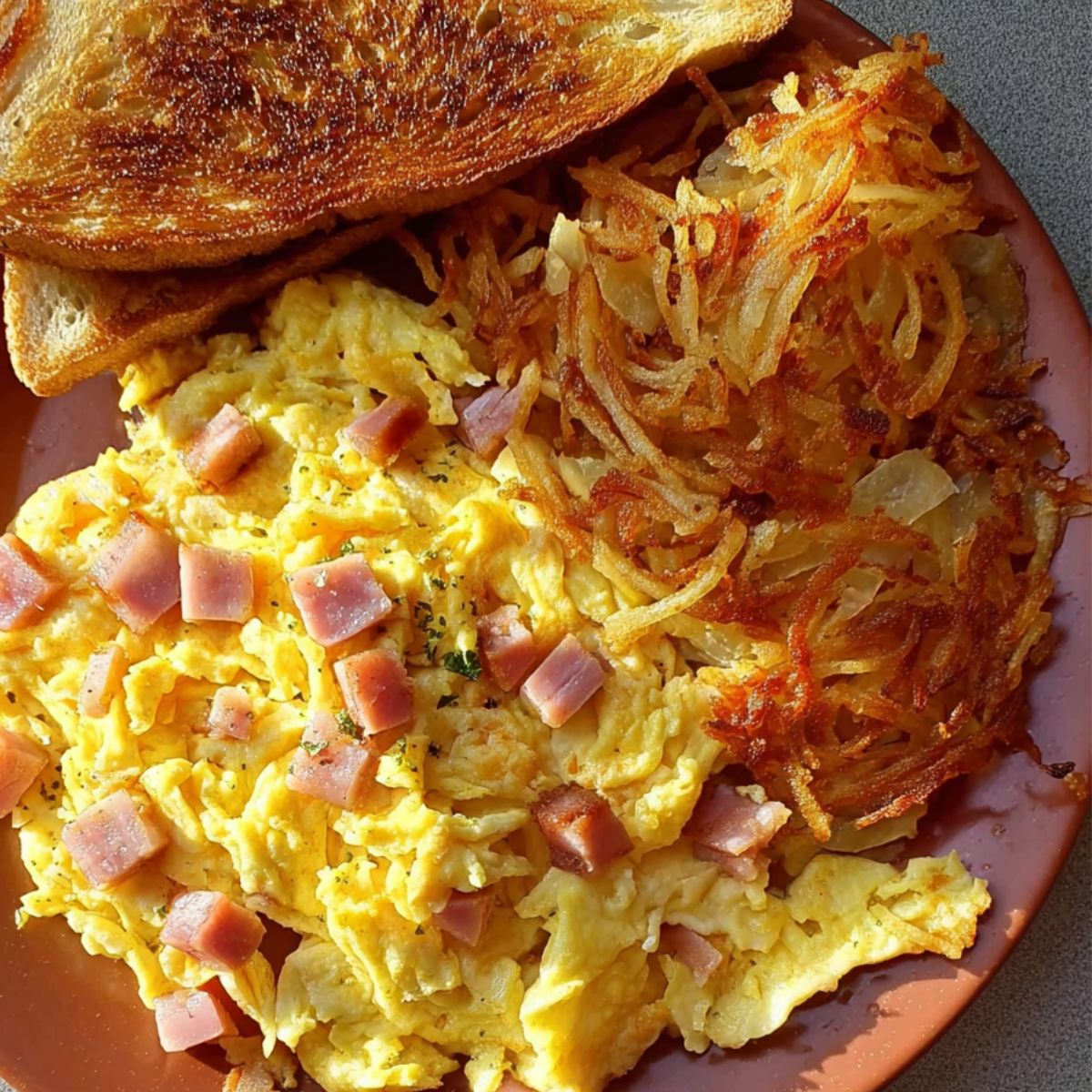 crispy hash browns A plate of scrambled eggs with diced ham, served with crispy hash browns and a slice of toasted bread.