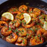 peri peri shrimp Cooked shrimp in a skillet, coated in a rich, reddish-brown sauce and garnished with chopped parsley and two lemon slices, ready to serve.