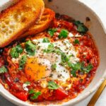 Finished shakshuka served in a bowl with a poached egg on top, garnished with fresh herbs and accompanied by toasted bread.