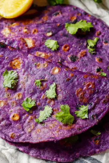 Easy Sweet Potato Tortillas Recipe 1 sweet potato tortillas Top-down close-up of a vibrant purple tortilla on a striped cloth, garnished with fresh cilantro, with a lemon wedge and a small bowl of chopped herbs beside it. The tortilla has charred spots, showing it’s freshly cooked, on a neutral textured surface. Bright, natural lighting emphasizes the rich color and texture of the tortilla, ideal for healthy wrap, vegan tortilla, or gluten-free recipe content.