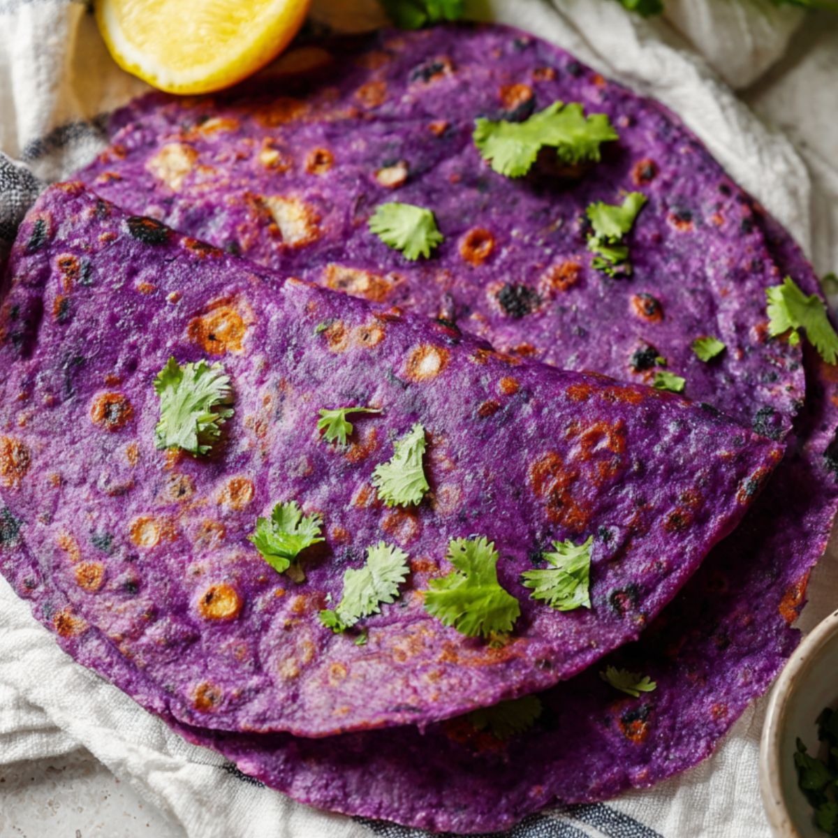 sweet potato tortillas Top-down close-up of a vibrant purple tortilla on a striped cloth, garnished with fresh cilantro, with a lemon wedge and a small bowl of chopped herbs beside it. The tortilla has charred spots, showing it’s freshly cooked, on a neutral textured surface. Bright, natural lighting emphasizes the rich color and texture of the tortilla, ideal for healthy wrap, vegan tortilla, or gluten-free recipe content.