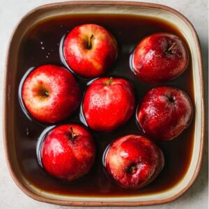 The Best Baked Peaches Recipe 16 Whole red apples placed in a dish and submerged in a dark brown liquid, possibly a sauce or syrup.