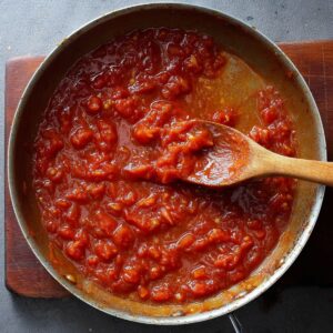 Healthy Blackened Shrimp Pasta Recipe 16 A pan containing a thick tomato sauce with small chunks of tomatoes, being stirred with a wooden spoon.