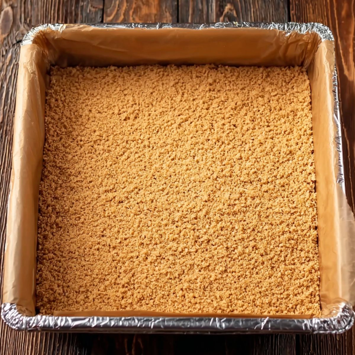 Healthy Butterfinger Caramel Crunch Bars Recipe 10 A pan lined with foil or parchment with a pressed layer of a crumbly base, likely made from crushed graham crackers, cookies, or a combination of crumbs and butter.