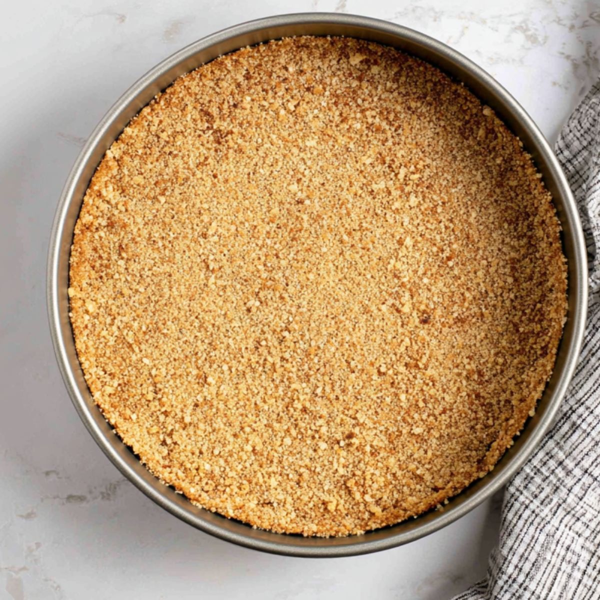 The Best Caramel Apple Cheesecake Recipe 11 "Overhead shot of a springform pan with an evenly pressed nut and graham cracker crust for caramel apple cheesecake, with a checkered kitchen towel nearby."