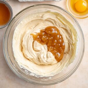 The Best Caramel Apple Cheesecake Recipe 16 "Top-down view of a glass mixing bowl with creamy cheesecake batter, including yogurt, applesauce, and caramel, next to a small bowl with an egg and a tray of eggs, ready for mixing."