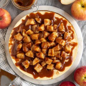 The Best Caramel Apple Cheesecake Recipe 17 "Overhead view of a creamy caramel apple cheesecake topped with cinnamon-spiced apple chunks and rich caramel sauce, surrounded by whole and halved apples, a jar of caramel sauce, a cake server, and a checkered kitchen towel."