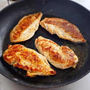 Healthy Chicken Asparagus Pasta Recipe 16 Four pieces of golden-brown, pan-seared chicken breasts cooking in a black skillet, with a slightly crispy surface.