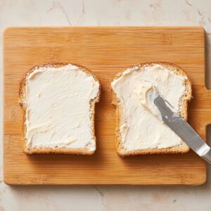 Two slices of bread on a wooden cutting board, spread with creamy mayonnaise using a butter knife, top-down view, natural lighting, clean and minimalistic sandwich prep