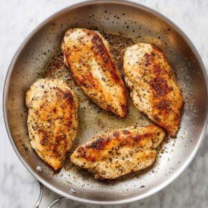 Four golden-brown chicken breasts cooking in a stainless steel skillet, seasoned with salt and pepper, top-down view, natural lighting, juicy and tender, home-cooked meal preparation.