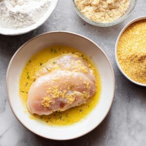 Easy Chicken Parmesan with Spaghetti Recipe 15 Raw chicken breasts coated in egg wash, with bowls of flour and seasoned breadcrumbs nearby for breading.