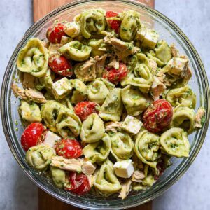 A glass bowl filled with green tortellini pasta salad mixed with shredded chicken, cherry tomatoes, and cubes of cheese, lightly coated in dressing.