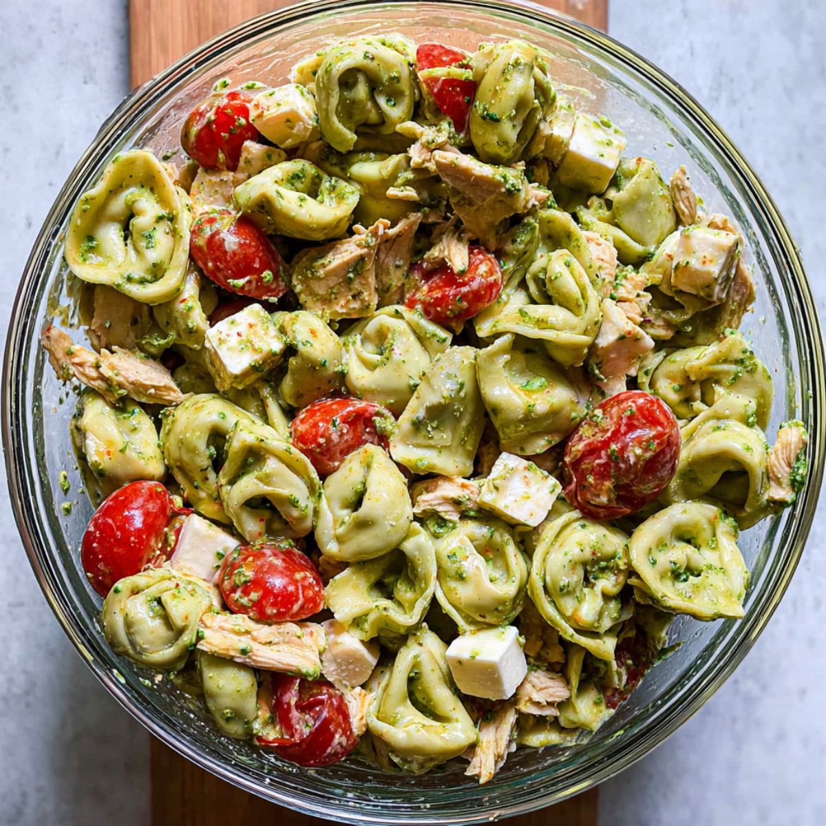 Best Chicken Tortellini Salad Recipe 12 A glass bowl filled with green tortellini pasta salad mixed with shredded chicken, cherry tomatoes, and cubes of cheese, lightly coated in dressing.