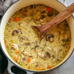 Delicious Chicken Wild Rice Soup Recipe 15 Grey pot with creamy chicken wild rice soup, containing chunks of chicken, sliced mushrooms, carrots, and wild rice. The wooden spoon is submerged in the soup.