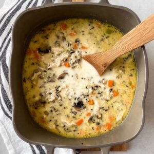 Delicious Chicken Wild Rice Soup Recipe 17 Top-down view of a creamy chicken wild rice soup in a grey rectangular pot, with chunks of chicken, mushrooms, carrots, and wild rice. A wooden spoon rests in the pot, and a black-and-white striped towel is visible in the corner
