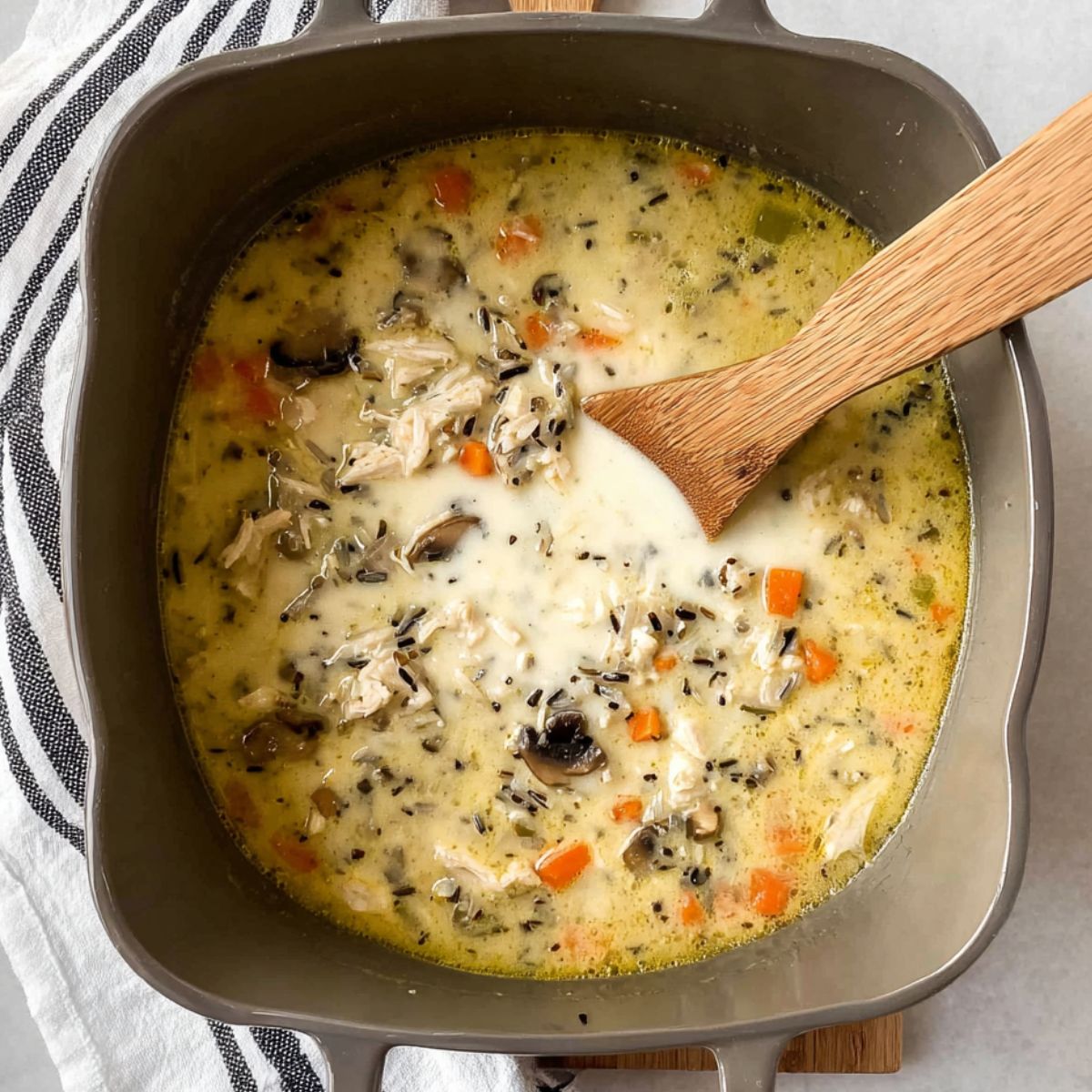 Delicious Chicken Wild Rice Soup Recipe 12 Top-down view of a creamy chicken wild rice soup in a grey rectangular pot, with chunks of chicken, mushrooms, carrots, and wild rice. A wooden spoon rests in the pot, and a black-and-white striped towel is visible in the corner