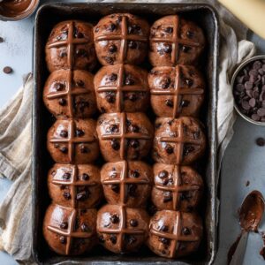 Healthy Chocolate Hot Cross Buns Recipe 17 A tray of freshly baked chocolate hot cross buns with chocolate crosses piped on top, ready to serve, with a piping bag and scattered chocolate chips nearby.